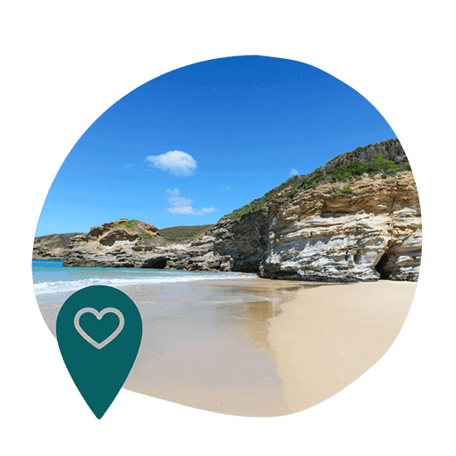 Serene beach with golden sands and rocky cliffs under a vibrant blue sky. A heart symbol on a location pin overlays the corner, suggesting a favorite spot.