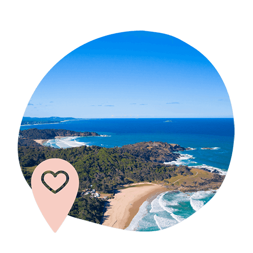 Aerial view of a serene coastal landscape with sandy beaches, lush greenery, and blue ocean under a clear sky. A pink location pin with a heart is overlayed.