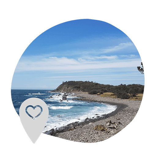 Rocky coastline under a bright blue sky with gentle waves lapping the shore, framed by a circular black border. A heart-shaped location pin overlays the image.