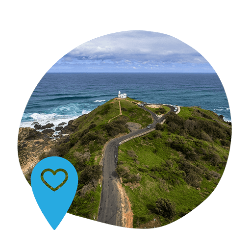 Aerial view of a winding road leading to a lighthouse on a green hill, surrounded by rocky coastline and ocean waves, under a cloudy sky. A blue map pin with a heart symbol is in the bottom left, adding a sense of location and affection.
