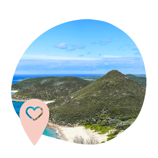 A scenic view of a lush green mountain by a beach with turquoise waters under a blue sky. A pink location pin with a heart symbol appears in the foreground.